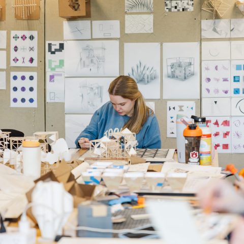 A person works on architectural models at a cluttered desk in a studio, with sketches, diagrams, and models displayed on the wall behind.