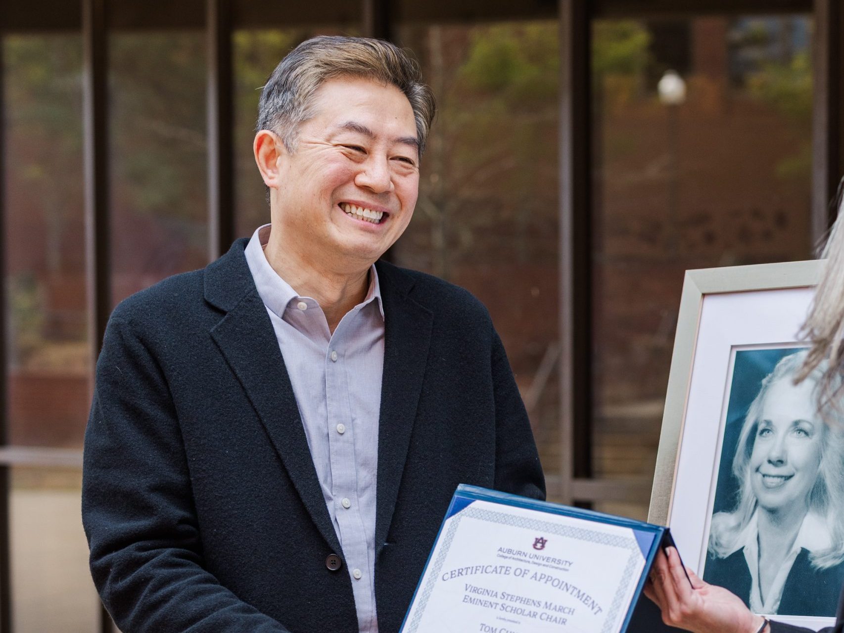 A man in a suit jacket smiles while receiving a framed certificate from another person; a framed photograph is displayed on an easel nearby.