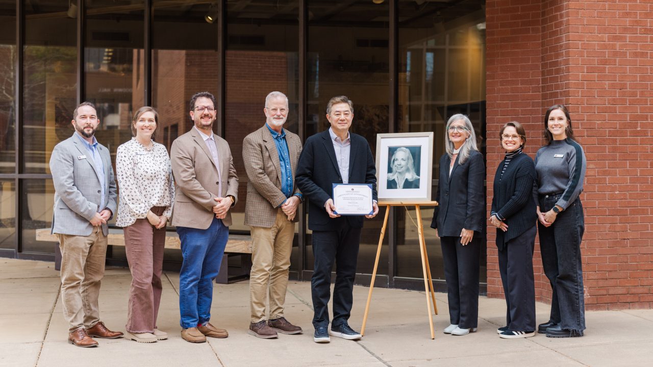 Eight people stand in a line outdoors near a building; one person holds a certificate next to a framed portrait on an easel.