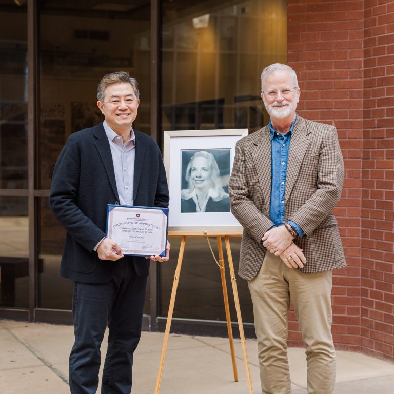 Two men stand beside a framed black-and-white portrait on an easel; one holds a certificate. They are outside a building with glass and brick walls.