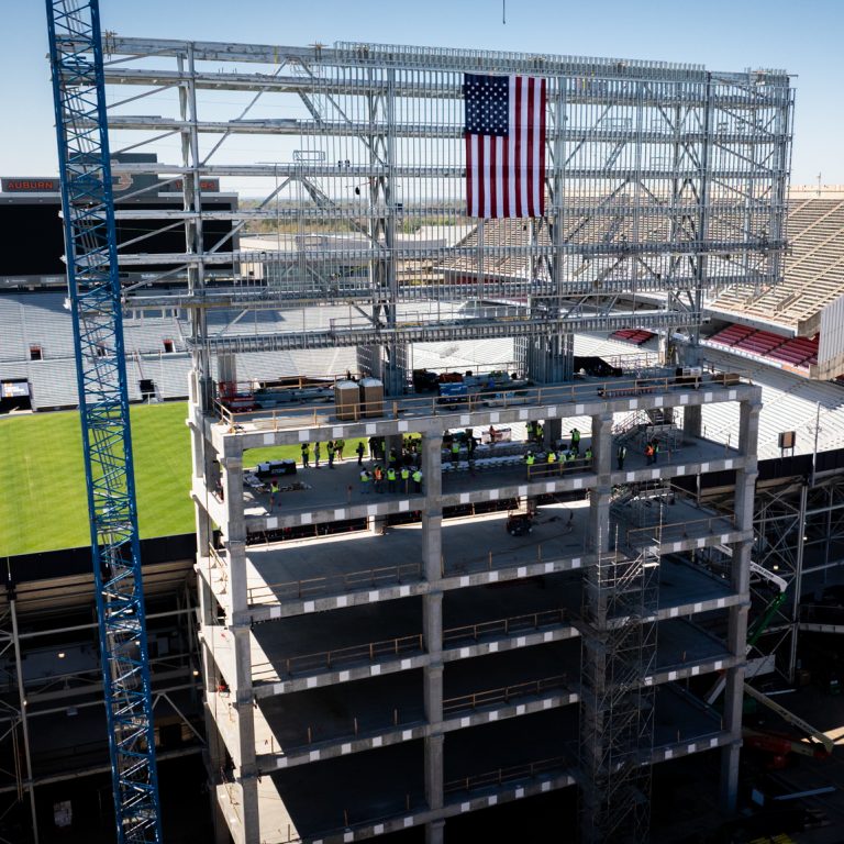 A steel stadium structure under construction with a large American flag hanging from the upper framework, adjacent to a partially completed seating area and a green field.