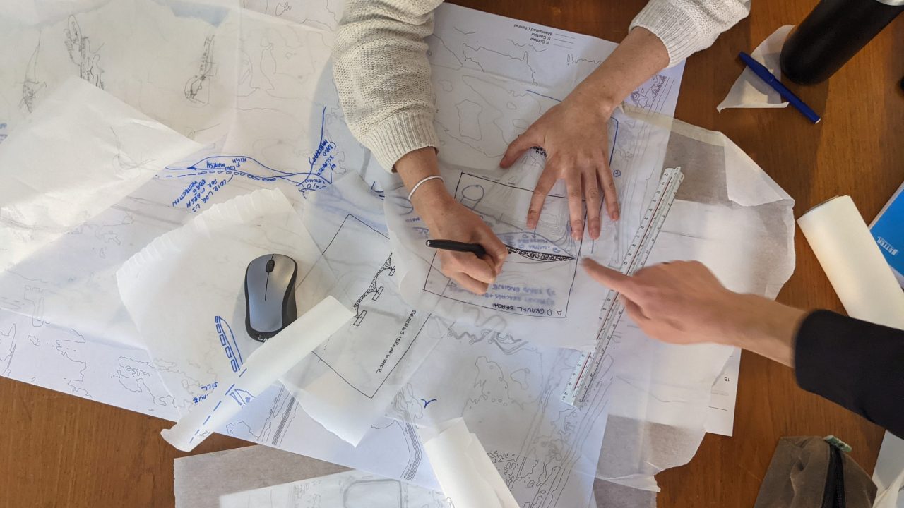 Two people working at a table covered with sketches, tracing paper, a ruler, a mouse, and drawing tools, collaborating on a design project.