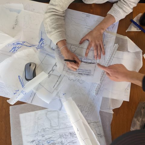 Two people working at a table covered with sketches, tracing paper, a ruler, a mouse, and drawing tools, collaborating on a design project.