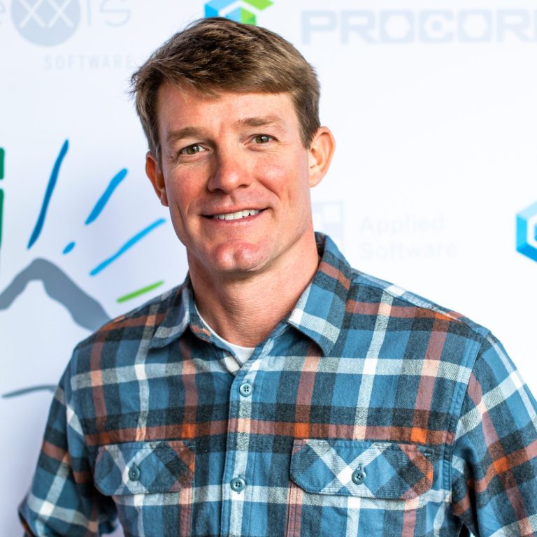 Man in blue, orange, and white plaid shirt smiles against a white backdrop with logos.