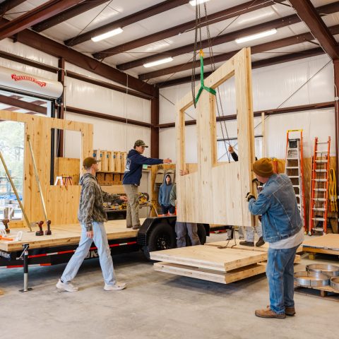 Five people assembling large wooden wall panels on a trailer inside a metal workshop; a crane is lifting one panel into place.
