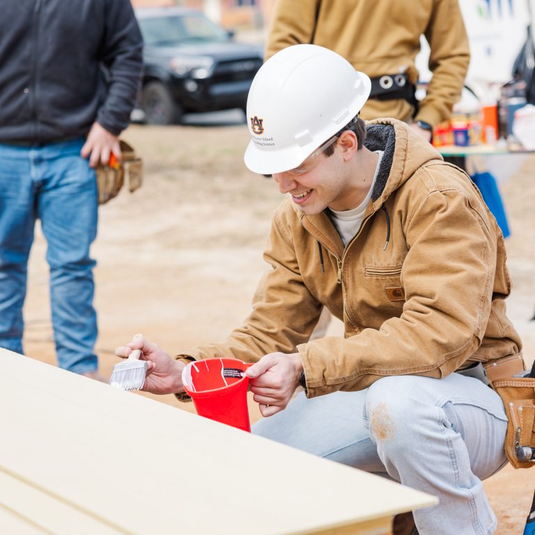 A man in a hard hat and brown jacket kneels and uses a measuring tool on wood boards at a construction site, while others stand nearby smiling.
