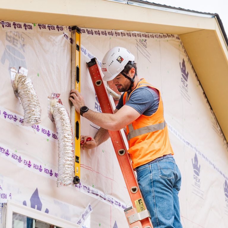 A construction worker wearing a hard hat and safety vest uses a level on the exterior wall of a house while standing on a ladder.