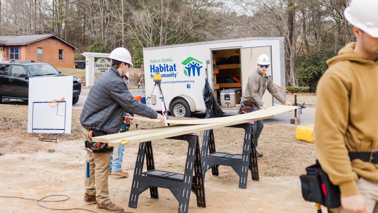 Several people wearing construction gear work with lumber outdoors at a Habitat for Humanity site, with a branded trailer and trees visible in the background.