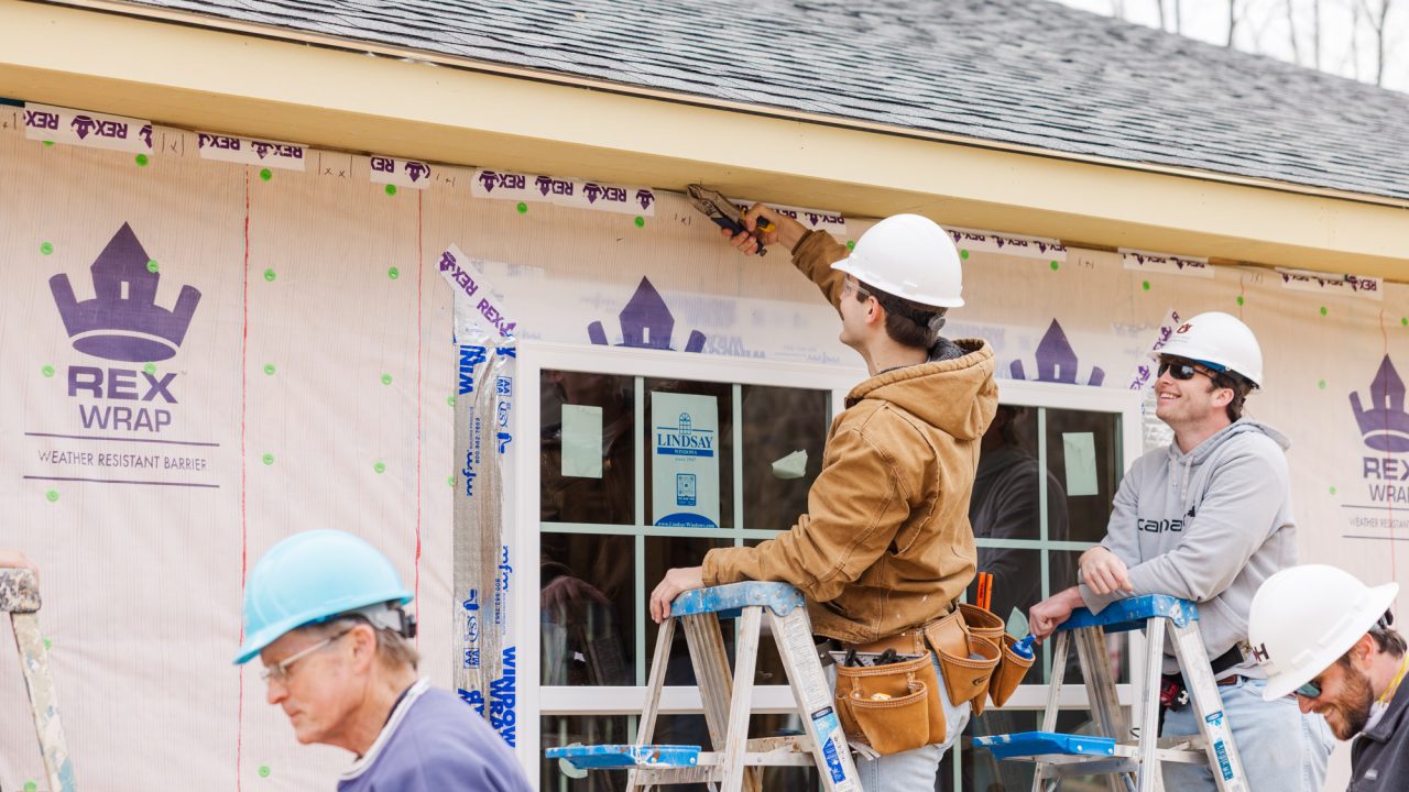 Four people wearing hard hats work on the exterior of a house under construction; two stand on ladders near a door, attaching material just below the roof.