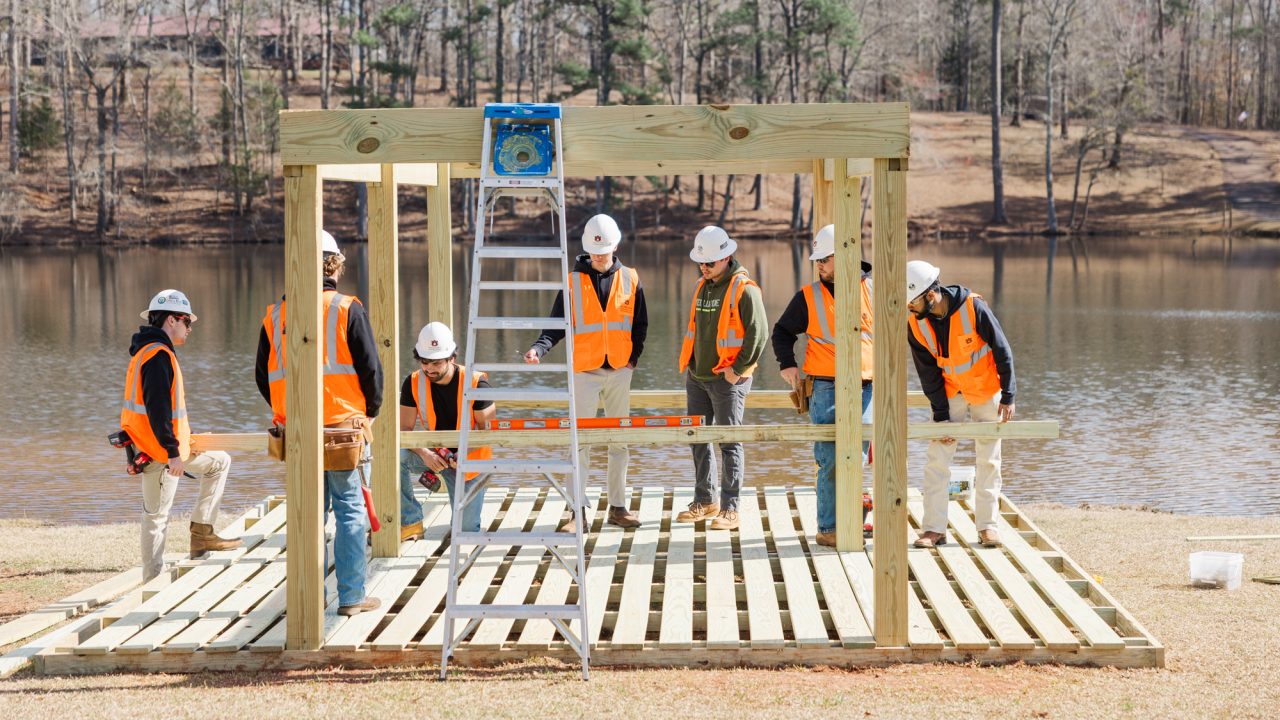 A group of construction workers in orange vests and hard hats assemble a wooden structure near a lake, with a ladder and tools in use.