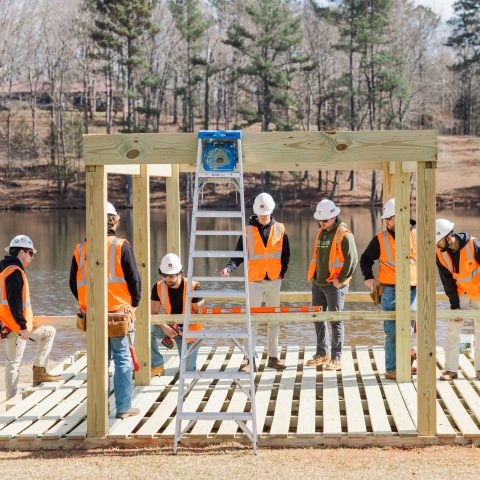 A group of construction workers in orange vests and hard hats assemble a wooden structure near a lake, with a ladder and tools in use.