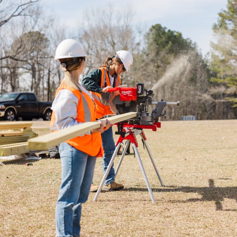 Two people wearing hard hats and orange safety vests operate a power saw to cut wood outdoors on a grassy field, with trees and vehicles in the background.