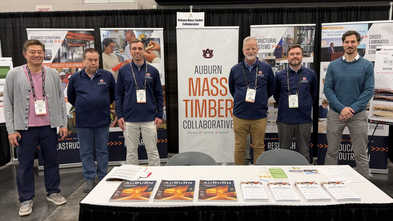 Six men stand behind a table displaying informational materials and magazines at the Auburn Mass Timber Collaborative booth during an indoor event or conference.