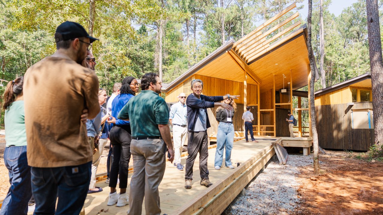 A group of people stands on a wooden walkway outside a modern cabin in the woods as a man in a suit gestures and speaks to them.