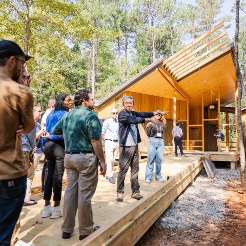 A group of people stands on a wooden walkway outside a modern cabin in the woods as a man in a suit gestures and speaks to them.