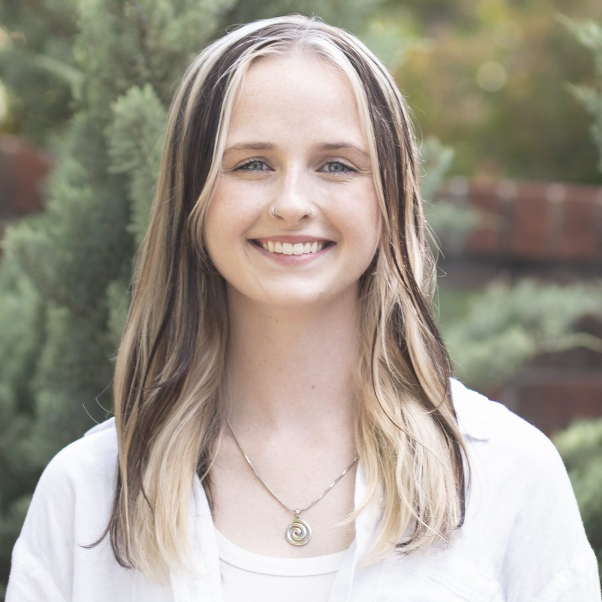 A young woman with straight, shoulder-length blonde and brown hair smiles outdoors, wearing a white shirt over a tank top and a pendant necklace. Green foliage and a brick wall are in the background.