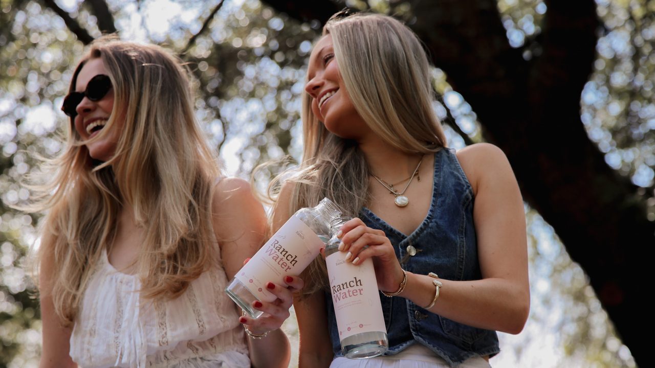 Two women outdoors holding bottles labeled Ranch Water, smiling and looking to the side, with trees blurred in the background.