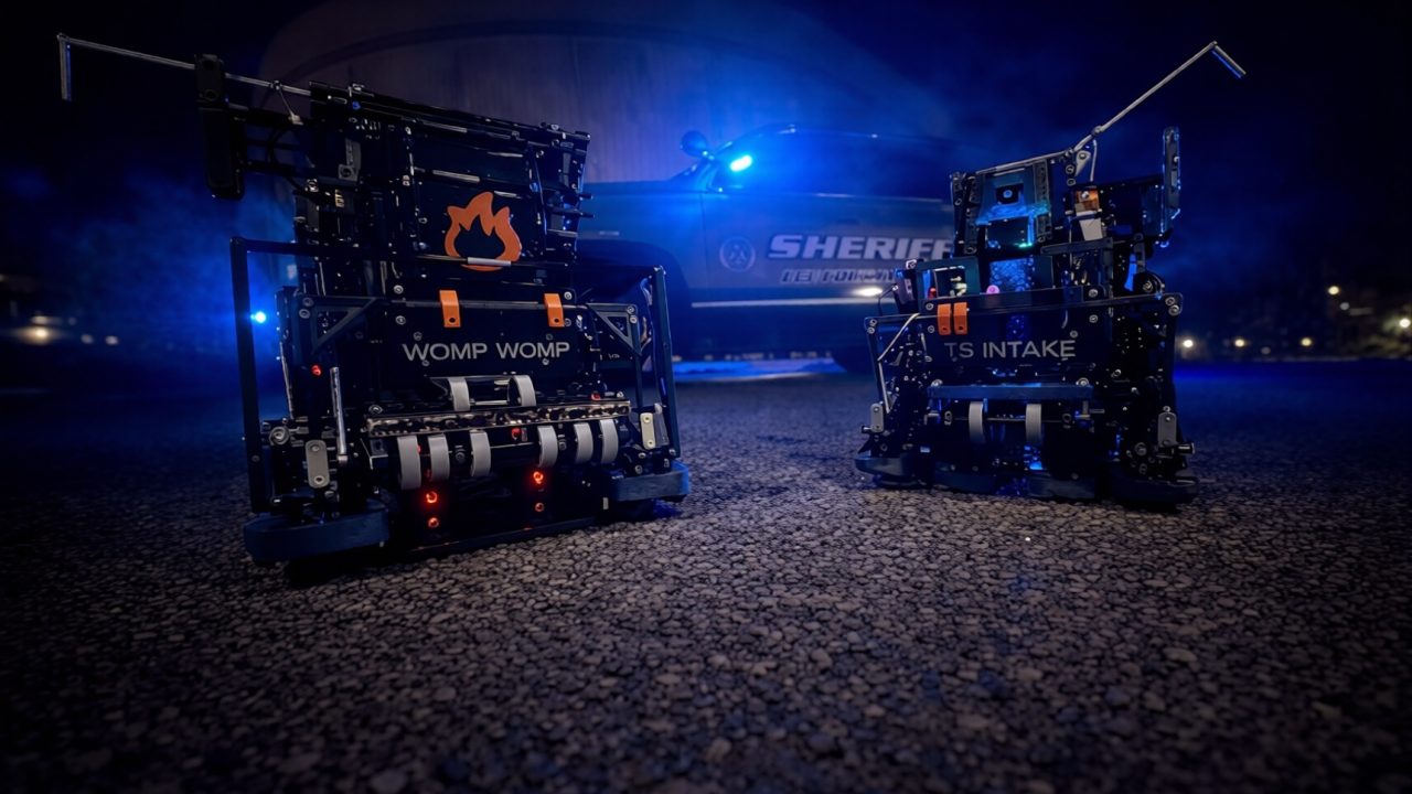 Two small robots labeled WOMP WOMP and TS INTAKE are positioned on gravel at night, with a police car and blue lights in the background.
