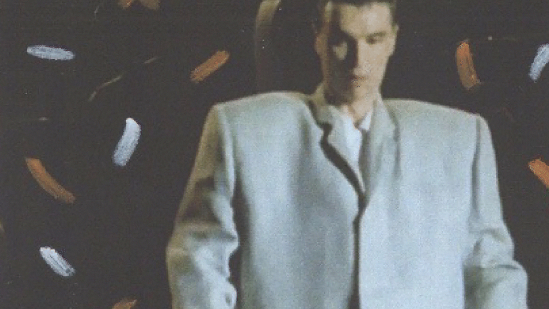 A person wearing an oversized gray suit sits against a dark background with scattered brushstroke patterns.