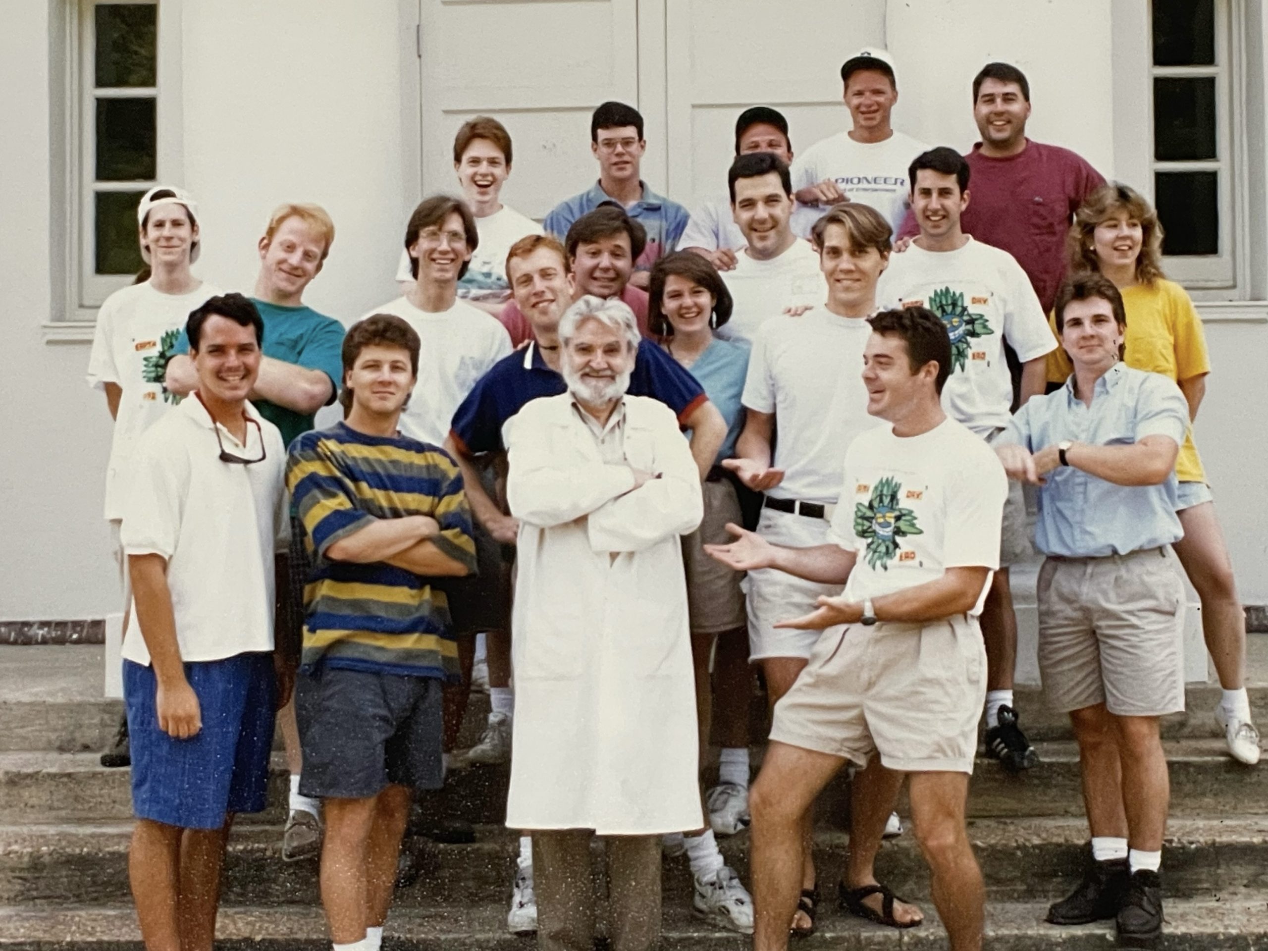 A group of young adults poses with an older man in a white coat on outdoor steps in front of a building, all smiling at the camera.