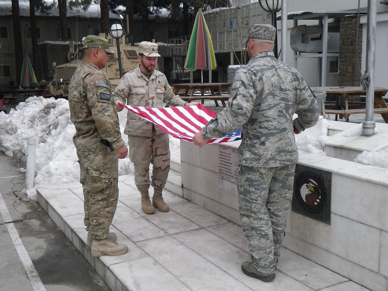 Three uniformed military personnel fold an American flag near a memorial with snow in the background.