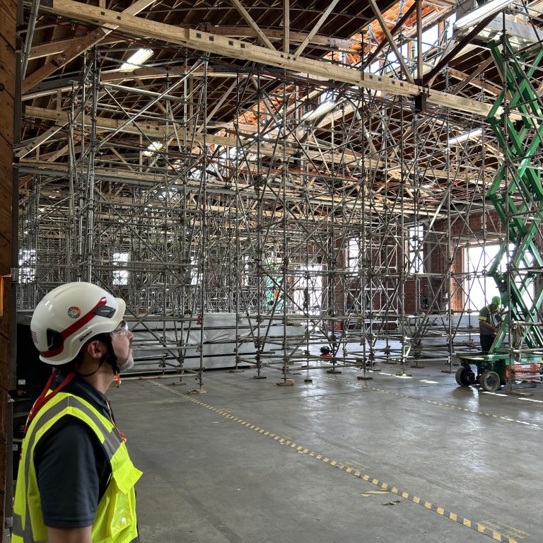 A worker in a hard hat and safety vest looks up at extensive scaffolding inside a large structure.