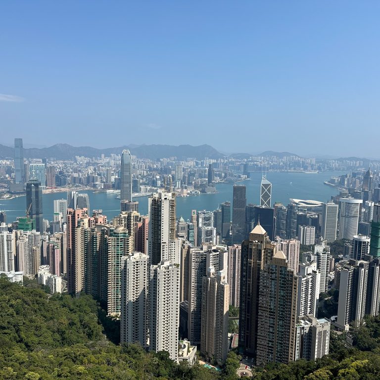 Aerial view of Hong Kong’s skyline with densely packed skyscrapers, Victoria Harbour, and mountains in the background under a clear blue sky.