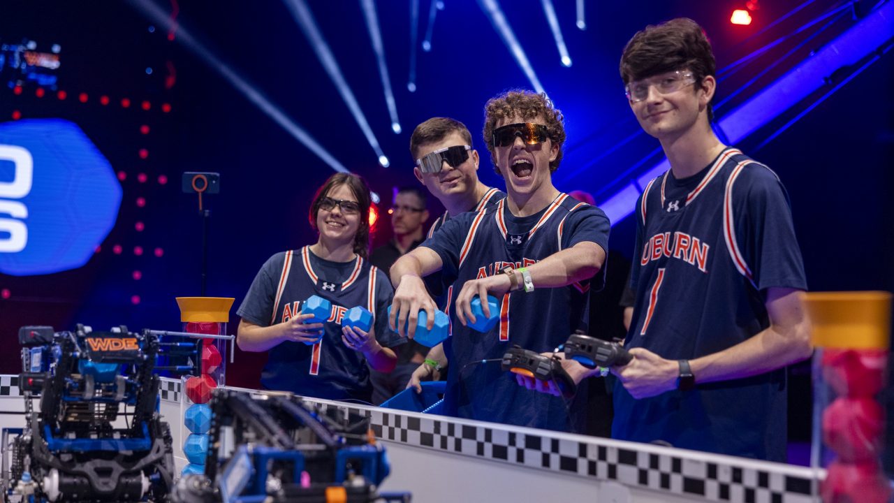 Four students in matching jerseys operate robots at a robotics competition, standing behind a barrier and controlling their devices with remote controllers.