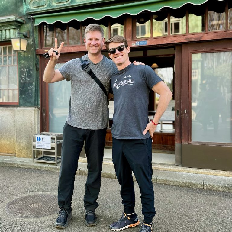 Two men stand smiling in front of a green, weathered building. One man gives a peace sign, both are casually dressed and wearing sunglasses.