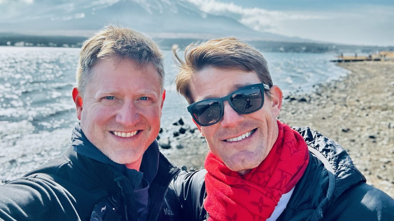 Two men smiling by a rocky lakeshore with a snow-capped mountain and partly cloudy sky in the background.