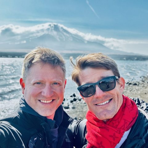 Two men smiling by a rocky lakeshore with a snow-capped mountain and partly cloudy sky in the background.