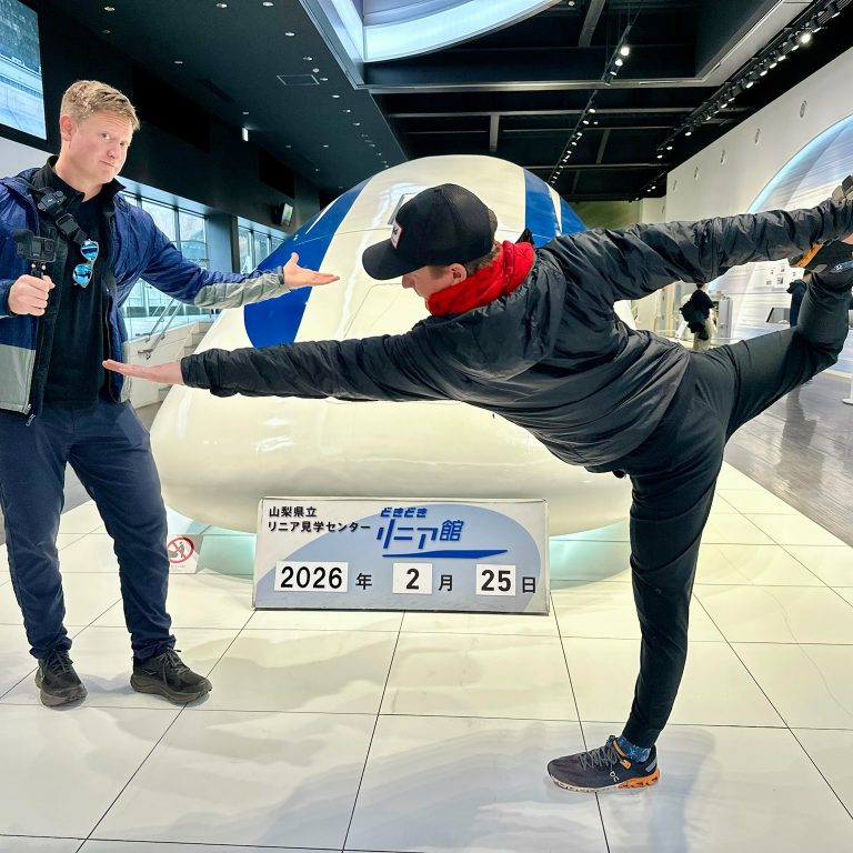 Two people pose playfully in front of a train nose display indoors; one stands holding a camera, the other balances on one leg with an arm outstretched.