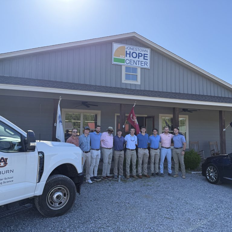 A group of people stands in front of the Jonestown Hope Center building beside a truck labeled Auburn McWhorter School of Building Science and a car, under a clear blue sky.