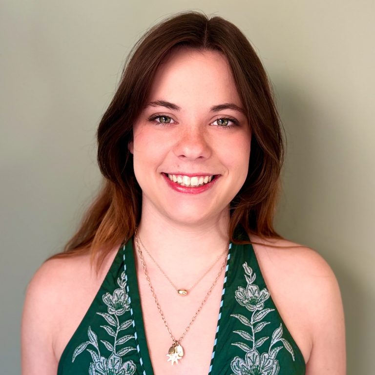A young woman with long brown hair, wearing a green floral top and layered necklaces, smiling in front of a plain light green background.