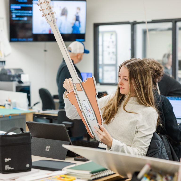 A woman sits at a desk in an office holding a large cardboard guitar cutout, with laptops, notebooks, and other office supplies around her.