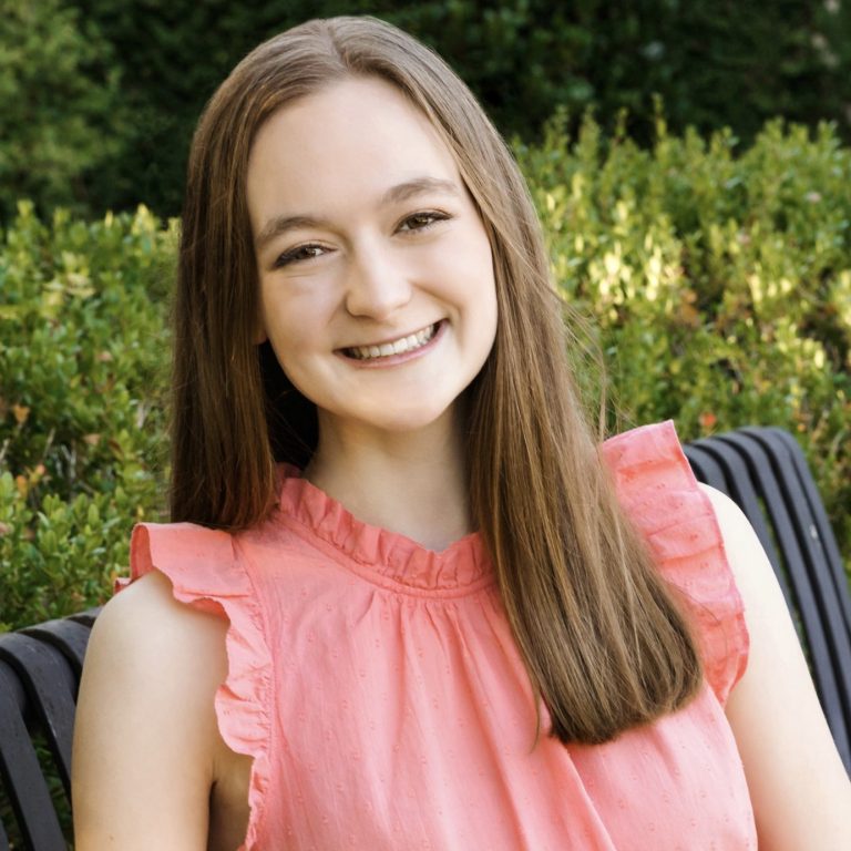 A young woman with long brown hair, wearing a sleeveless pink blouse and white pants, sits on a black metal bench outdoors in front of green bushes, smiling at the camera.