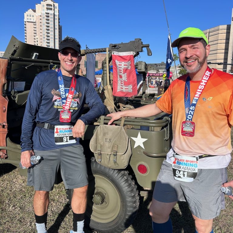 Two men wearing race finisher medals stand beside a vintage military jeep outdoors, with buildings and a blue sky in the background.