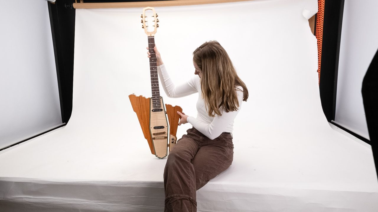 A person in a white shirt and brown pants sits on a white surface and holds a uniquely-shaped wooden guitar in a photo studio setting.