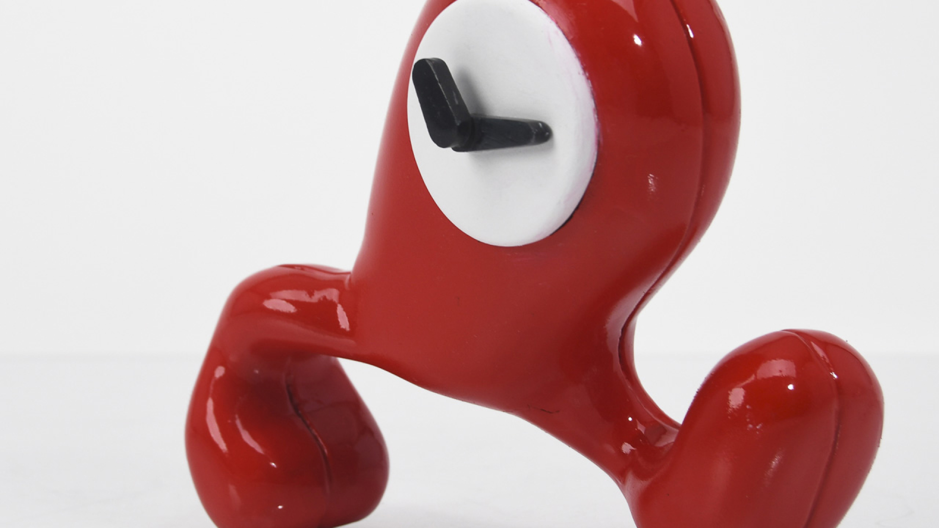 A red, abstract, three-legged figure with a white circular face and a black L-shaped object protruding from its center, against a plain white background.