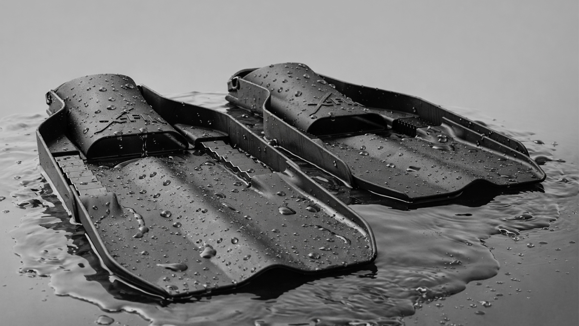 A pair of black swim fins with water droplets on them, resting on a reflective wet surface against a plain gray background.