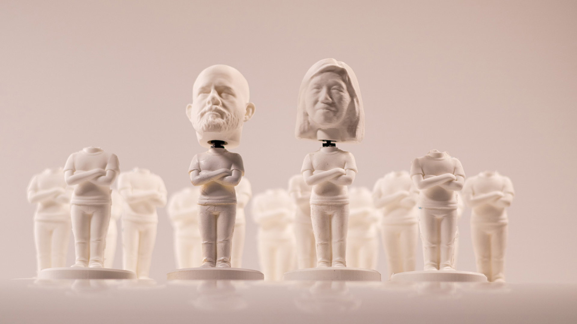 Rows of white figurines with folded arms, most headless, except two in front with detailed human heads, set against a plain background.