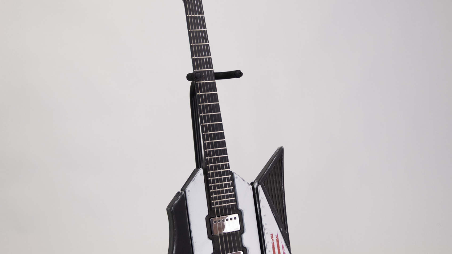 A uniquely shaped electric guitar with black and white panels, a US flag sticker, and metallic control knobs, standing on a black floor stand against a plain background.