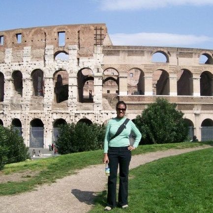 A person stands on a path in front of the Colosseum in Rome, Italy, on a sunny day. Shrubs and other visitors are visible nearby.
