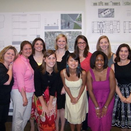 A group of ten women pose and smile in front of architectural plans and posters displayed on a wall.