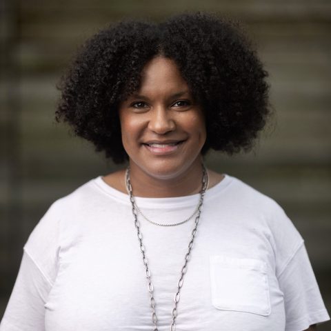 A woman with curly hair wears a white t-shirt and a long necklace, standing in front of a blurred, neutral background.