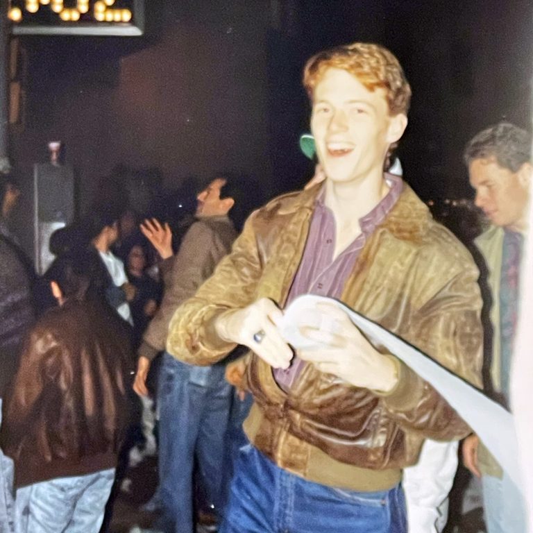 A young man in a brown jacket smiles and holds a roll of paper on a city street at night, with other people and scattered debris around him; a digital clock reads 5:02.