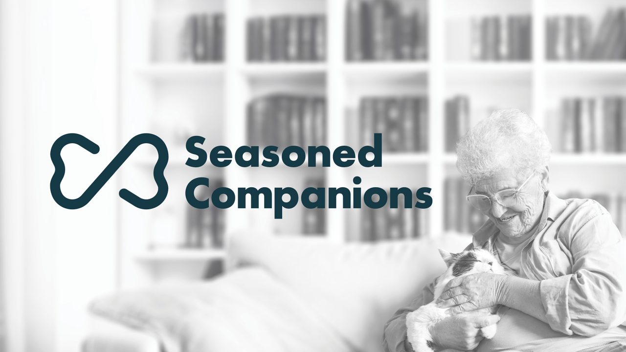 An elderly woman sits on a couch, smiling and petting a cat, with the Seasoned Companions logo and stylized bone shape displayed beside her.
