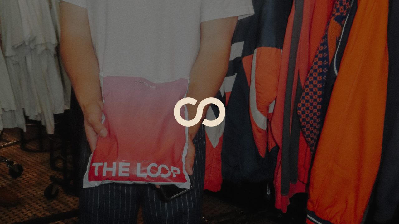 Person holding a red and white bag labeled THE LOOP in a clothing store, with racks of assorted garments visible in the background.