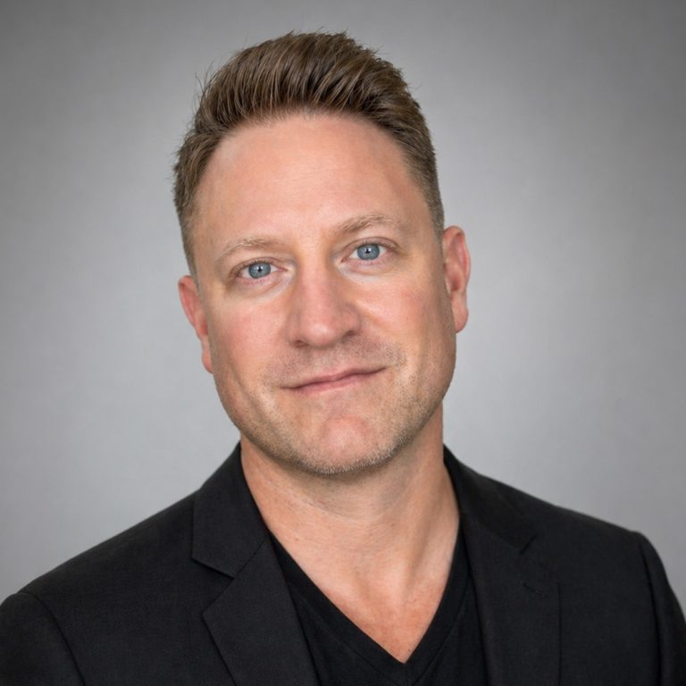 Man with short light brown hair wearing a black blazer and black shirt, facing the camera against a plain gray background.