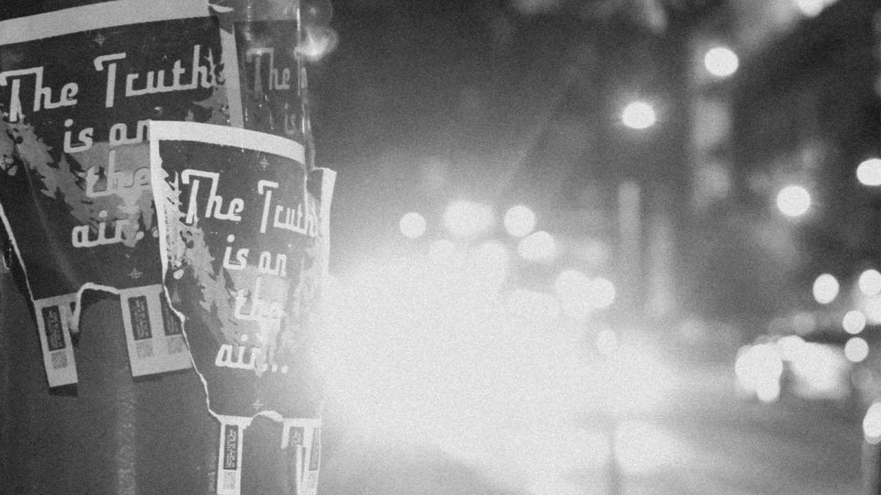 Black and white photo of torn posters on a pole reading The Truth is on the air, with blurry city lights and traffic in the background.